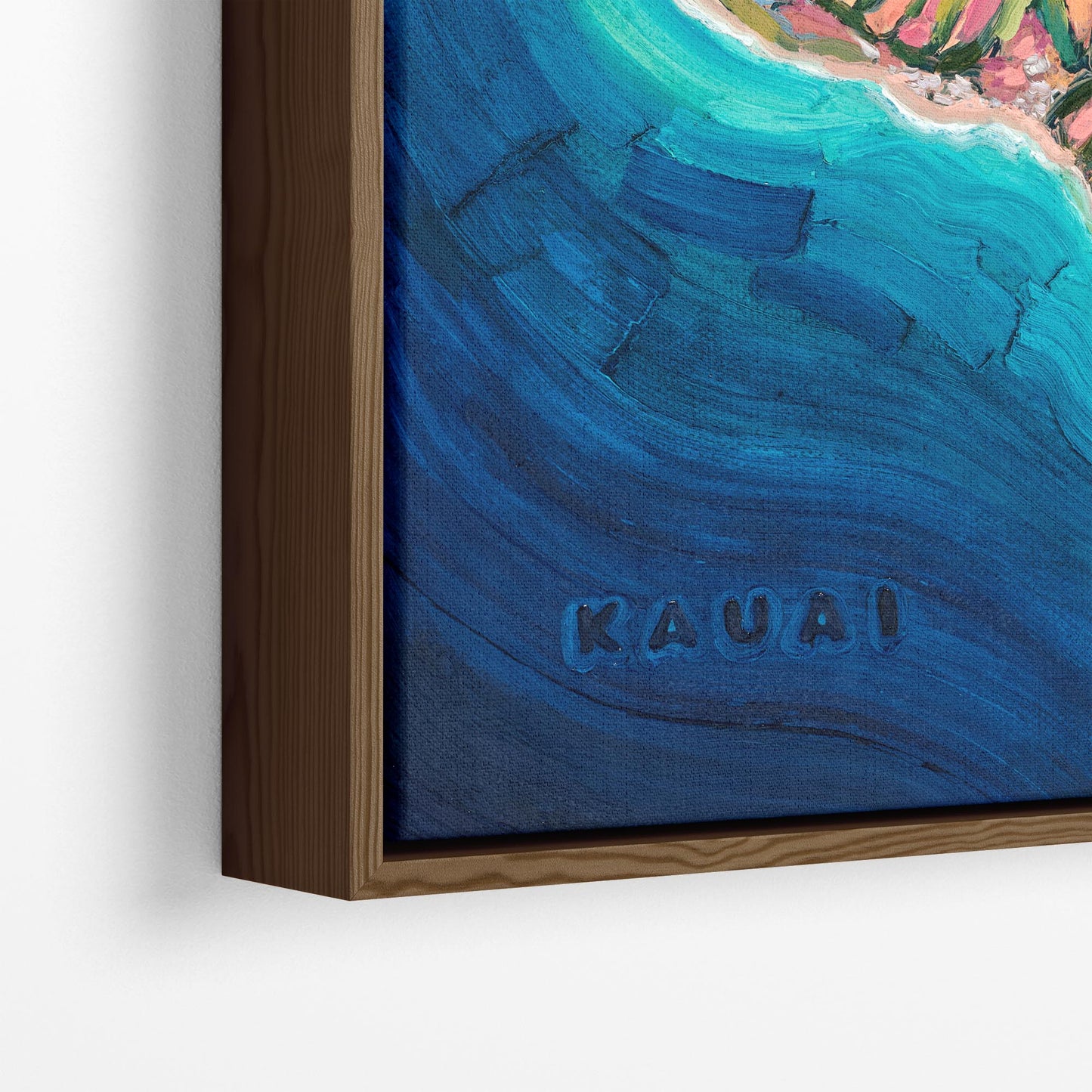 A close-up of a framed abstract coastal-style artwork showing deep blue water, turquoise tones, and a small section of pink shoreline, mounted on a white wall. The word "KAUAI" appears in the lower center of the artwork.