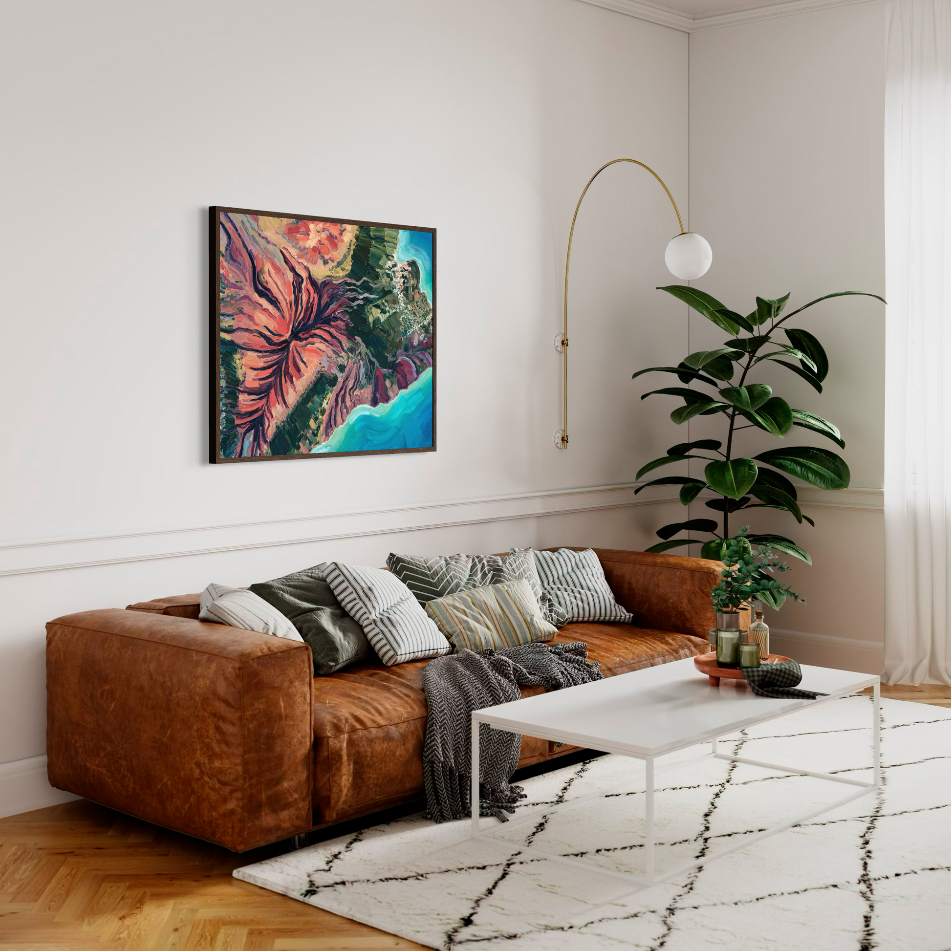 A styled living room featuring a large framed abstract painting with vibrant pink, orange, green, and blue tones hanging on a white wall above a brown leather sofa. The room includes a white coffee table, a white patterned rug, a tall green potted plant, and a gold floor lamp.