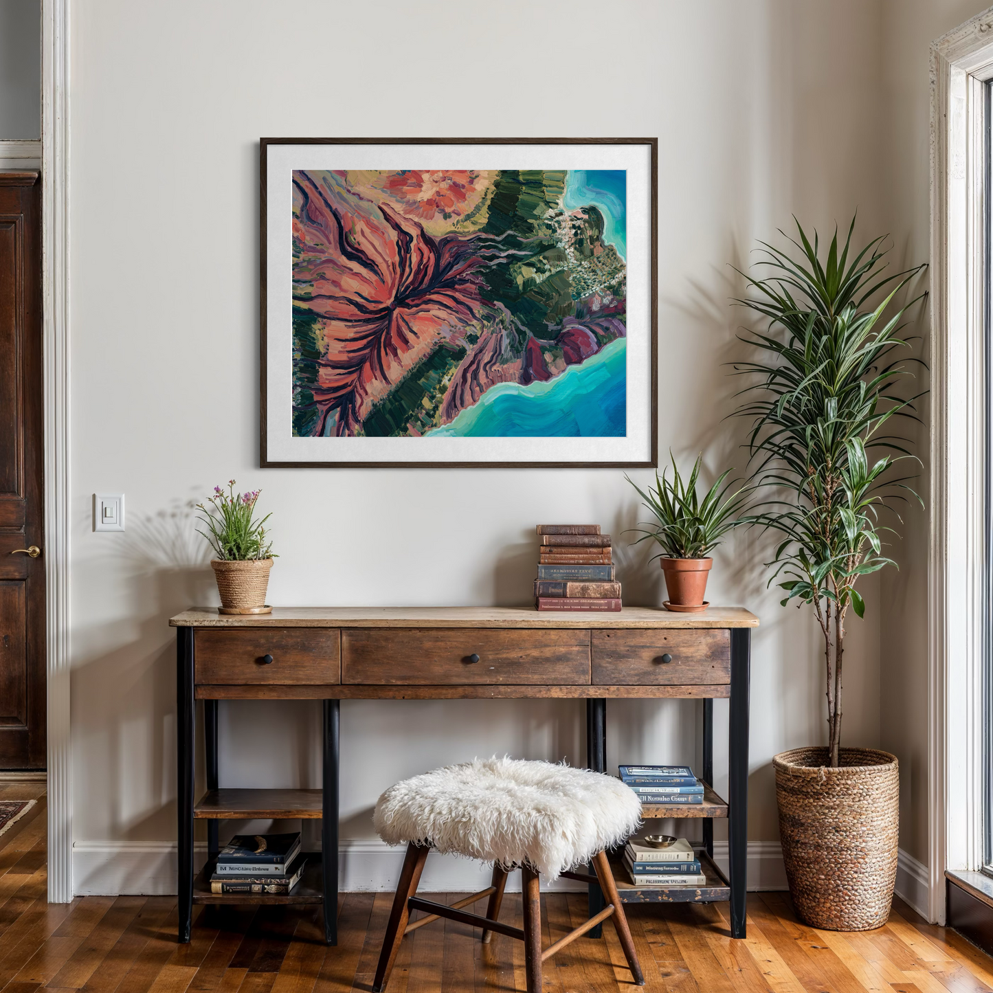 A framed abstract art print featuring vibrant red, green, and blue floral-like patterns, displayed on a beige wall above a wooden console table in a styled home office setting.