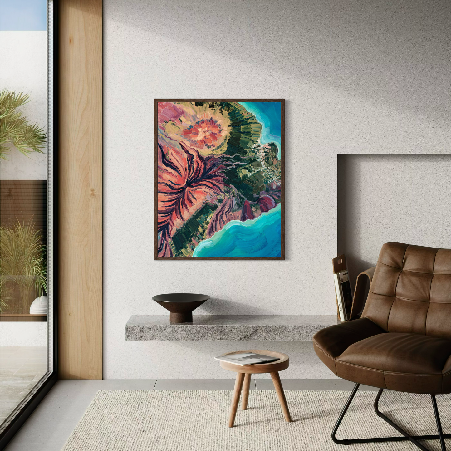 A colorful abstract framed wall art print hangs on a white wall in a modern living room setting with a brown leather armchair, a small wooden side table, and a stone shelf.