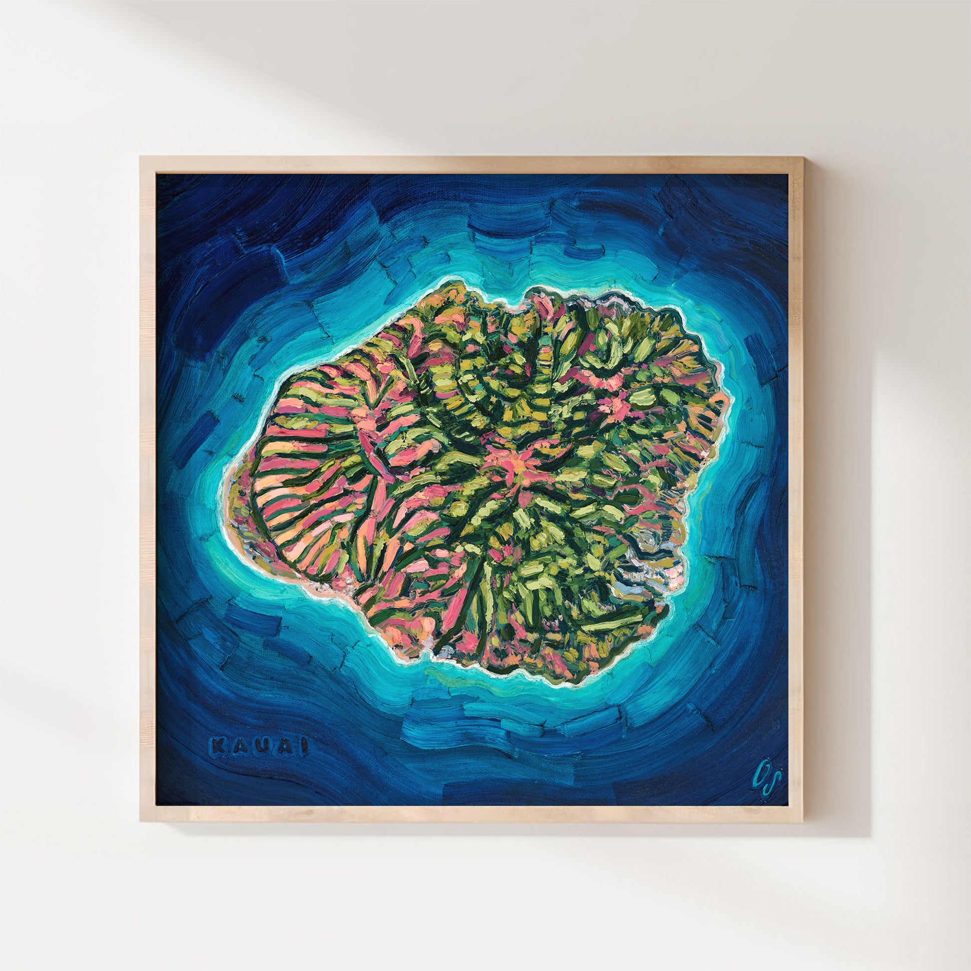 A framed square art print depicting a colorful coral reef island surrounded by deep blue and turquoise ocean water, hung on a light beige wall.